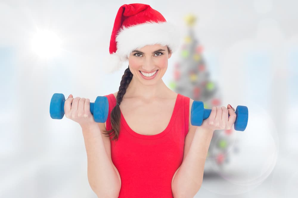 A woman in a Santa hat and red tank top lifting blue weights and showing off her weight loss results